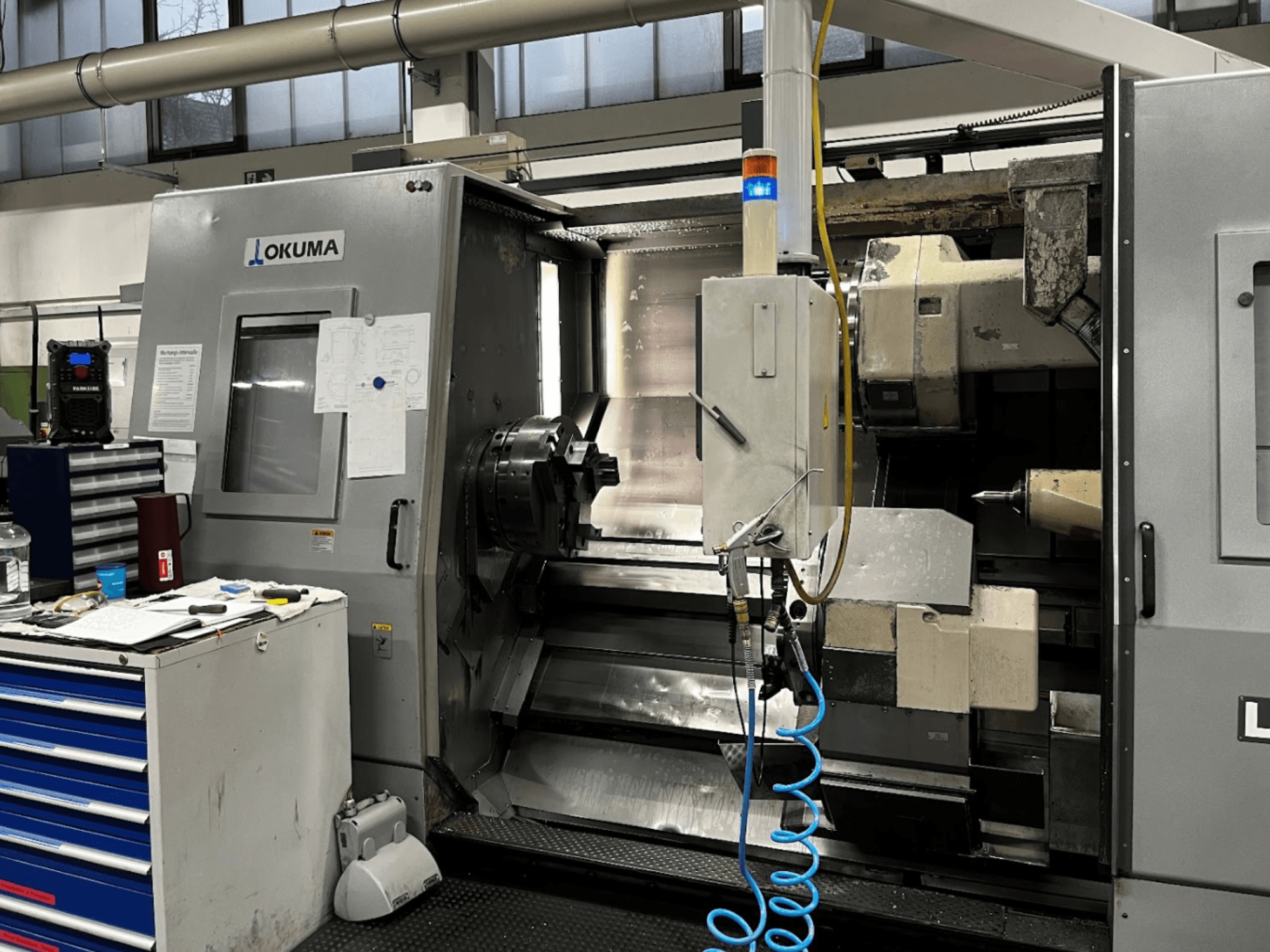 Torneiro horizontal Okuma LU35-M, vista frontal. Possui painel de controlo, ferramentas e mandril grande para operações de maquinagem.