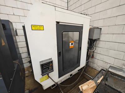 Máquina CNC industrial, vista lateral; apresenta painel de controlo, sinais de aviso e peças de madeira numa superfície de trabalho.